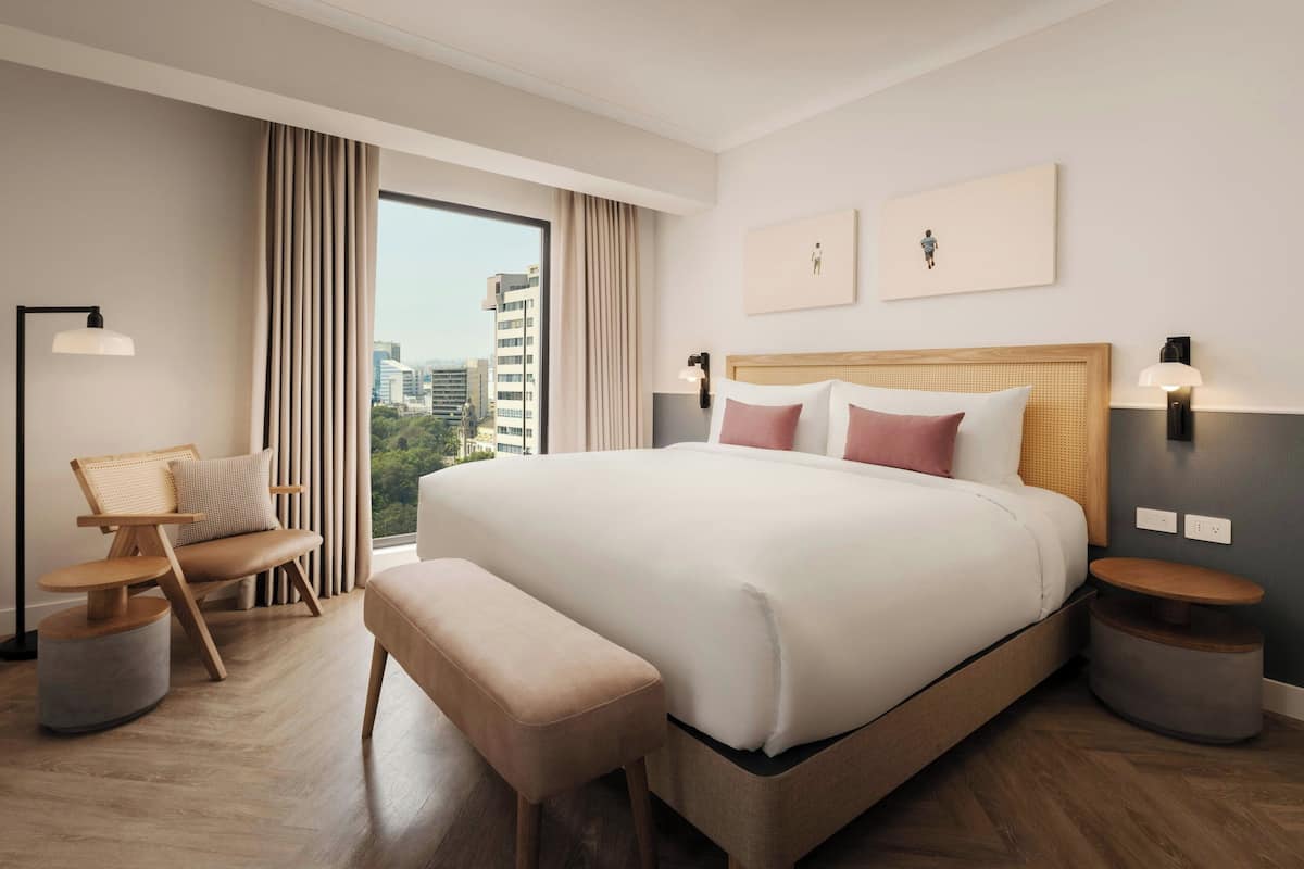 A modern hotel room with a large bed, a seating area with a chair and ottoman, a bedside table, and a view of the city through large windows.