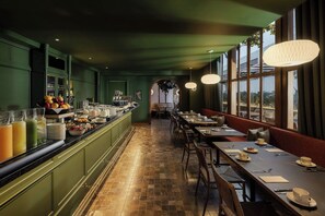 Uma longa área de refeições com um buffet de um lado, uma área de estar com mesas e cadeiras e grandes janelas do outro.