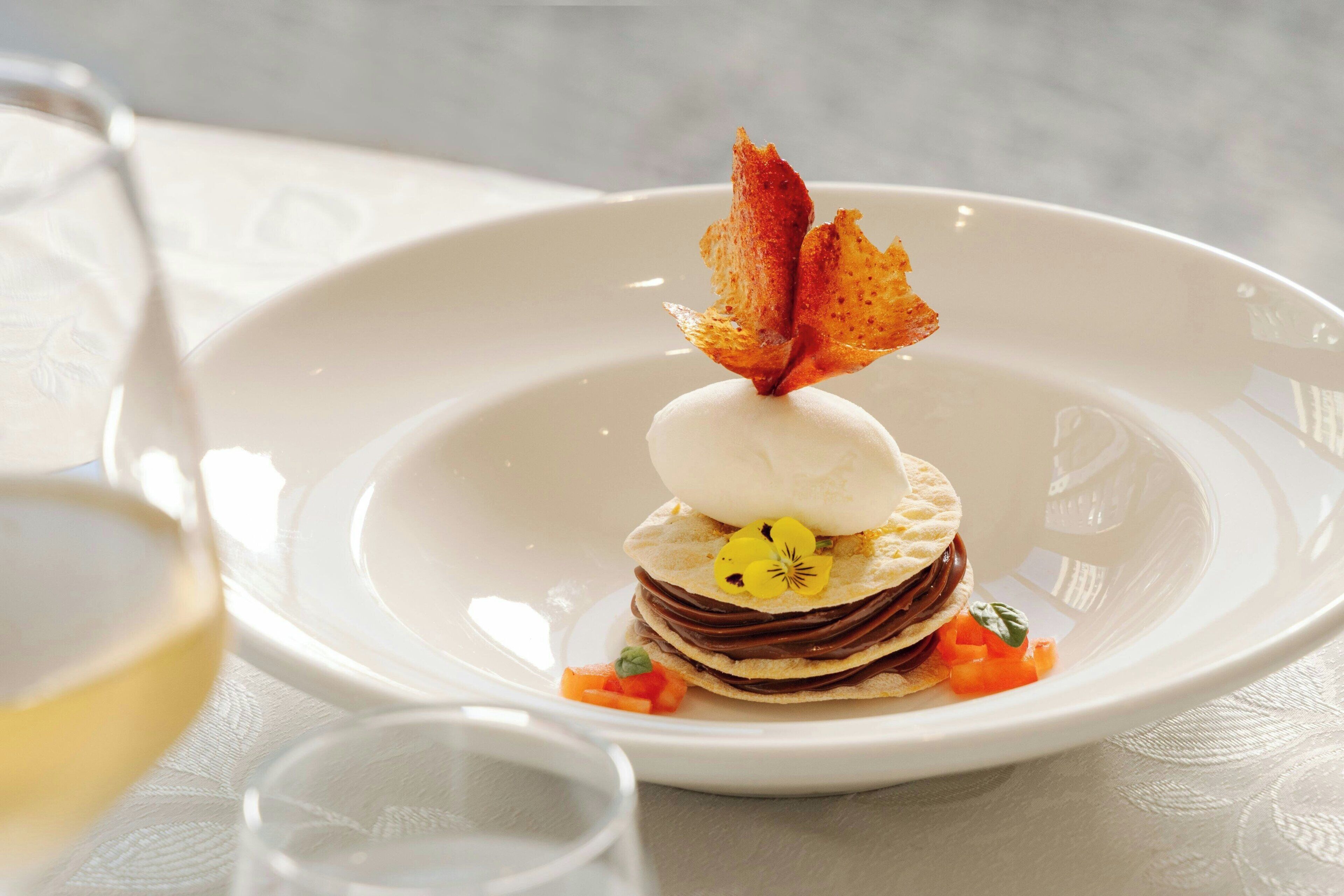 Un postre sobre un plato blanco, adornado con una hoja, una copa pequeña de vino blanco y un vaso de agua.