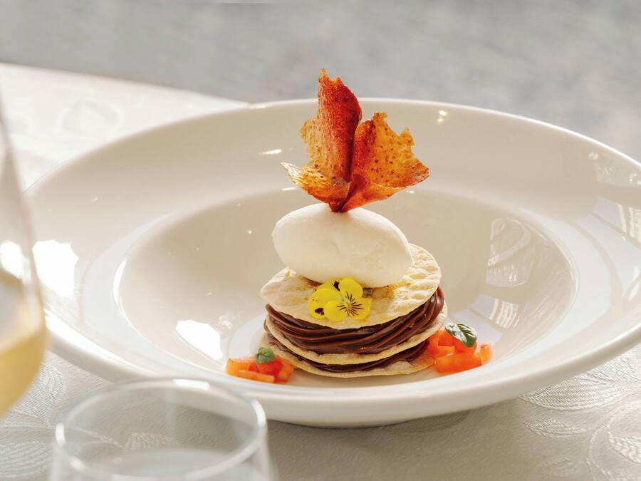 Un postre sobre un plato blanco, adornado con una hoja, una copa pequeña de vino blanco y un vaso de agua.