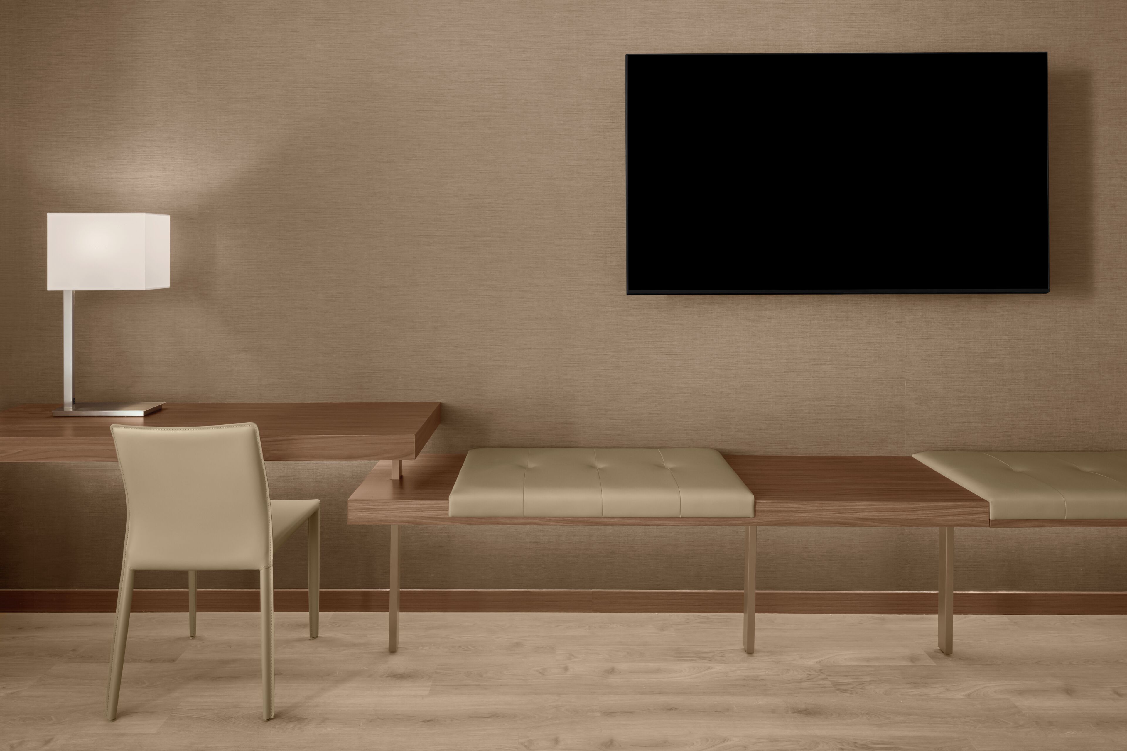 A modern hotel room with a wooden desk, a cushioned bench, a white chair, and a mounted TV.
