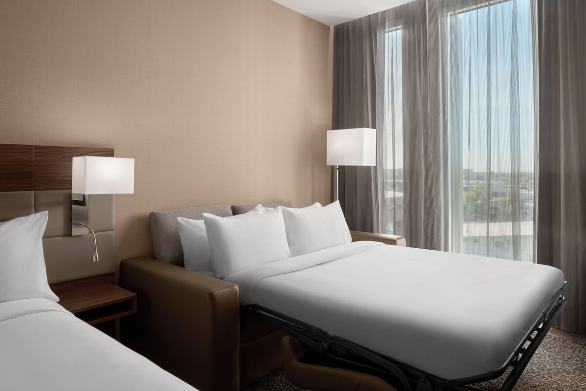 A hotel room with a bed, a sofa, a nightstand, a lamp, and a window with curtains.