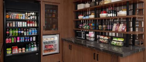Una cocina con refrigerador, microondas y estantes llenos de alimentos y bebidas.