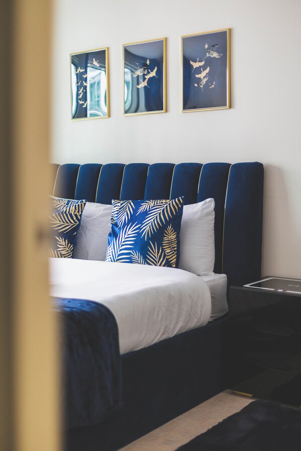 A bedroom with a dark blue upholstered headboard, a white bedspread, and patterned pillows. There are three framed artworks on the wall above the bed. - Deluxe Two-Bedroom Vauxhall Apartment (London)