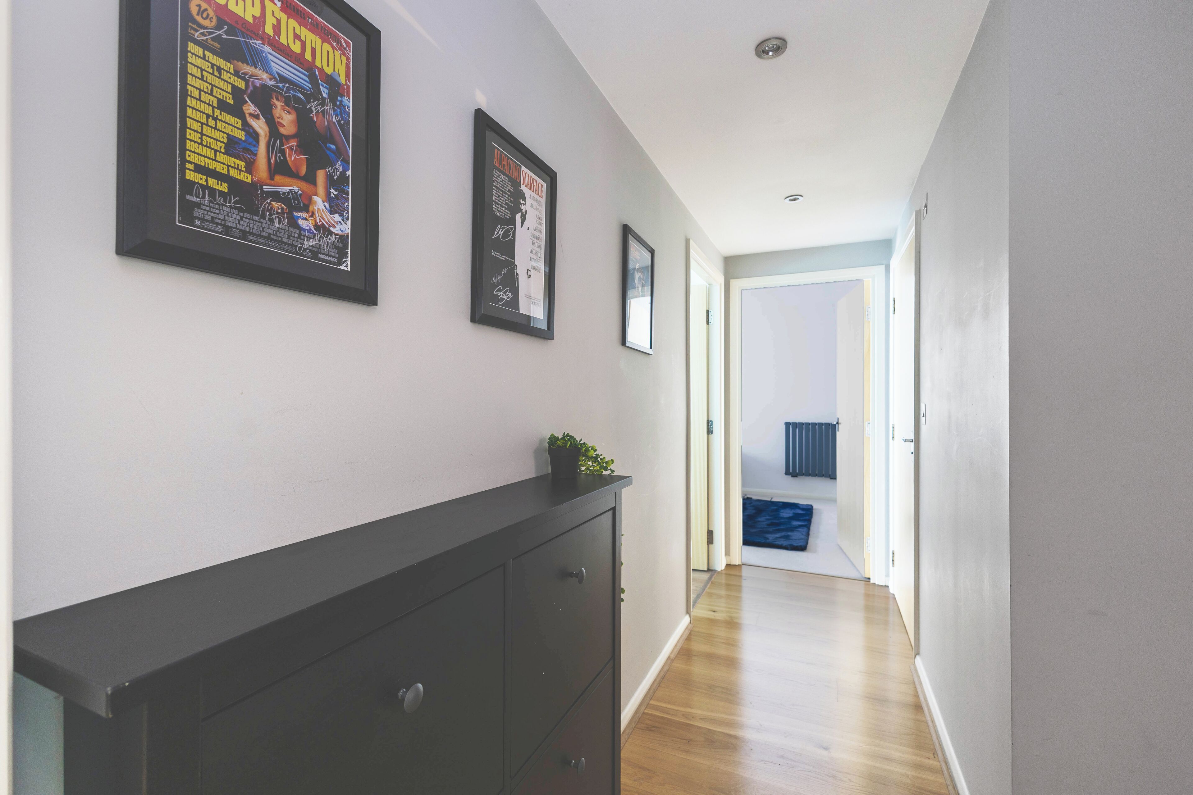 A hallway with framed posters on the wall, a dark-colored dresser, and a blue object on the floor.