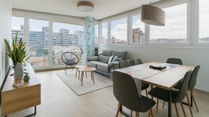 A modern living room with a sofa, dining table, and chairs, large windows with a city view, and a hanging light fixture. - 3BR Ocean Sunset Suite Las Canteras (Las Palmas de Gran Canaria)