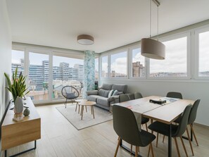 Ein modernes Wohnzimmer mit Sofa, Esstisch, Stühlen, großen Fenstern mit Stadtblick und einer Pendelleuchte.