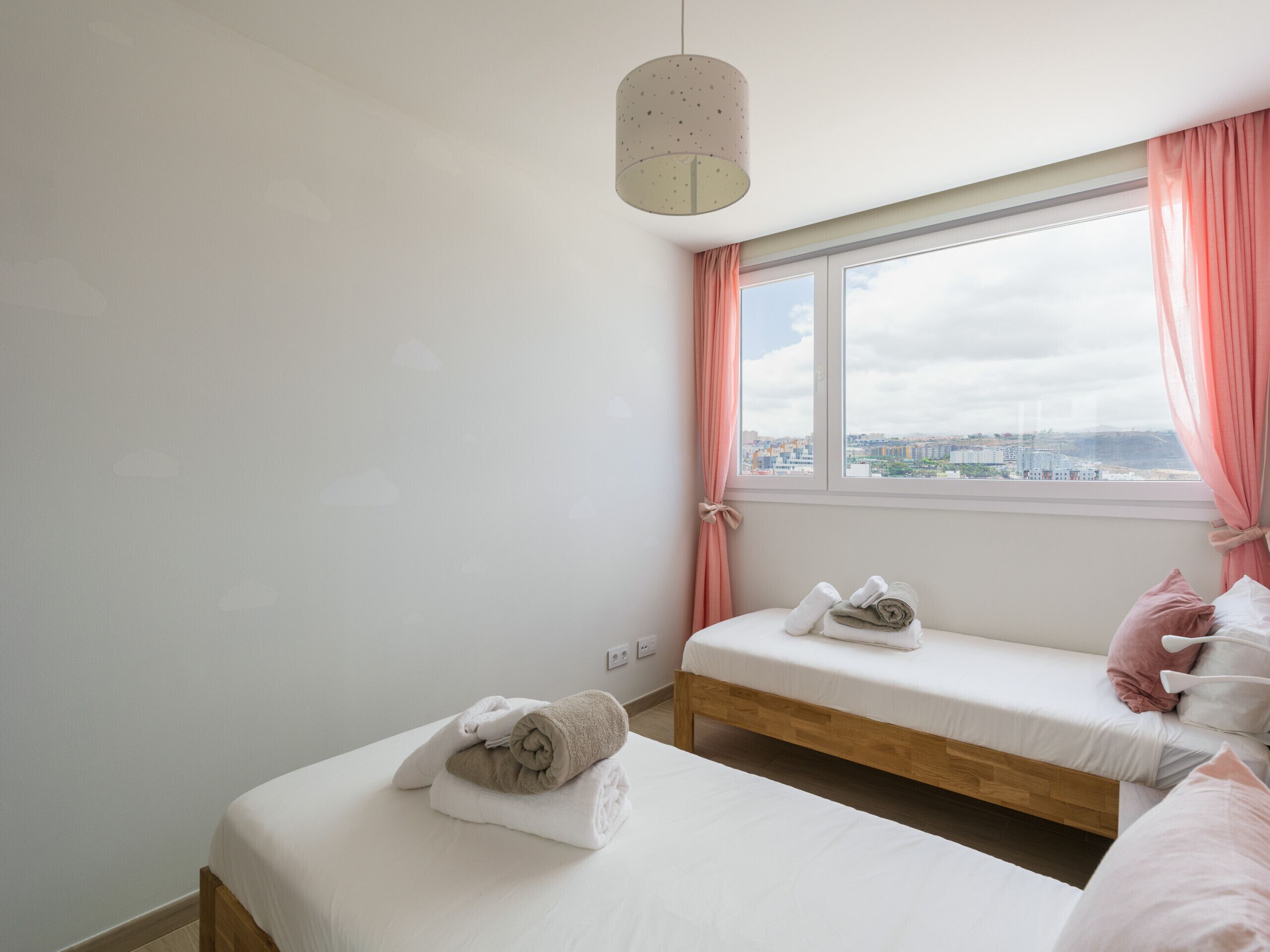 A bedroom with two beds, a window with pink curtains, and a city view.