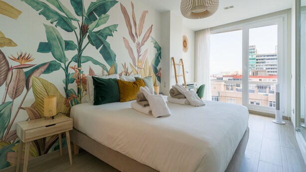 A bedroom with a large bed, a nightstand, a wall mural, a hanging light, and a view of buildings through a large window. - 3BR Ocean Sunset Suite Las Canteras (Las Palmas de Gran Canaria)