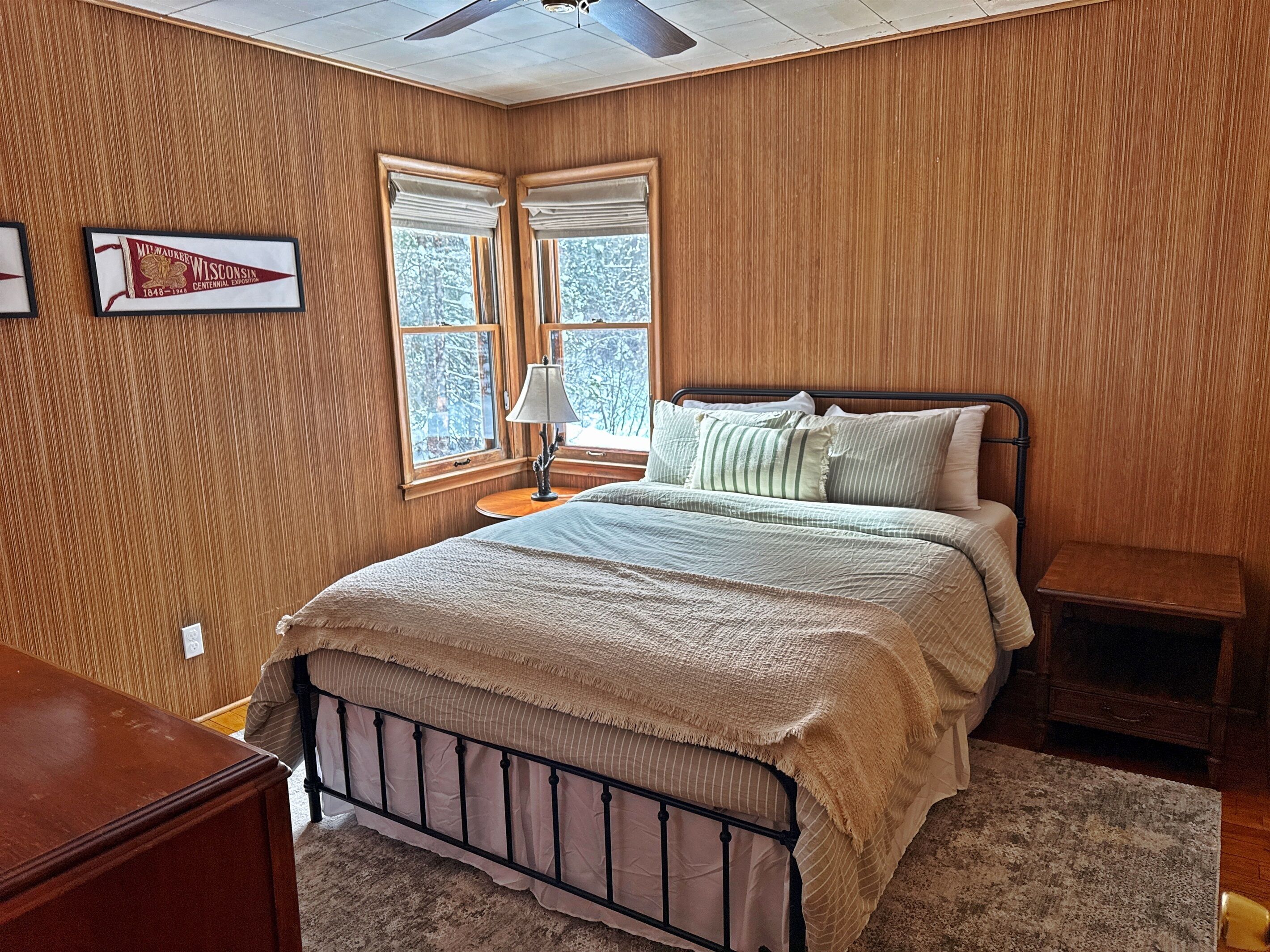 Een slaapkamer met een bed, nachtkastjes, een houten wand en een raam met uitzicht op bomen.
