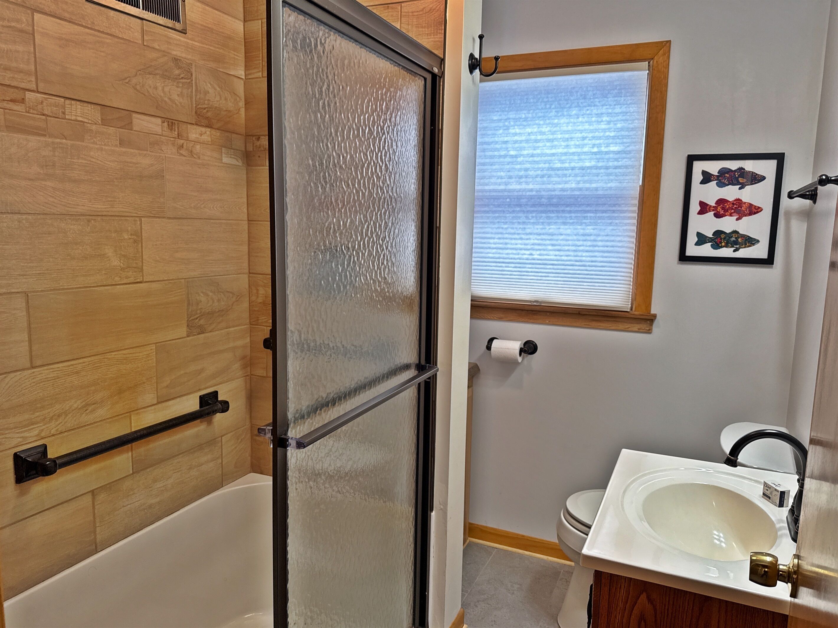 Un baño con pared de madera, puerta de ducha de vidrio, inodoro, lavabo y una obra de arte con peces enmarcados.