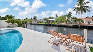 A swimming pool with two wooden lounge chairs, a clear view of a canal, and a residential area with palm trees and houses in the background. - Intracoastal Villa | Pool + 40ft Dock | Sleeps 8 (Fort Lauderdale)