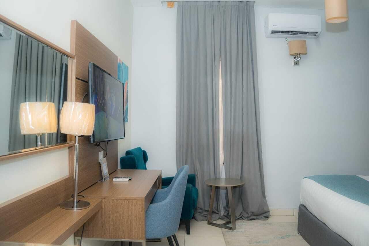 Une chambre d’hôtel avec un bureau en bois, deux lampes, une télévision, un lit et une fenêtre avec des rideaux.