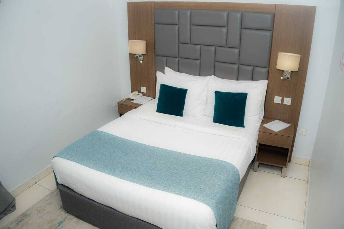 Una cama bien hecha con ropa de cama blanca y aguamarina, un cabecero tapizado en gris y dos mesitas de noche con lámparas.
