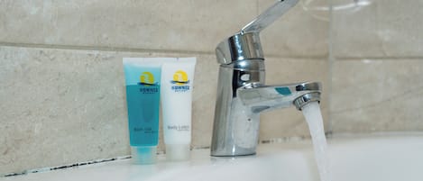 Un lavamanos con grifo cromado y agua corriendo, dos artículos de tocador con la etiqueta 'RowneZ' (uno azul y uno blanco), y una pared de azulejos color beige.