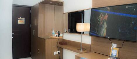 A hotel room with a wooden desk, a television mounted on the wall, a wardrobe, and a trash bin.