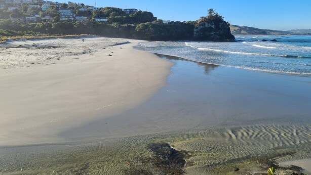 Una playa con agua cristalina, una orilla arenosa y un promontorio rocoso.