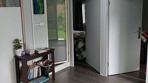 Una habitación con suelo de madera, una estantería con libros y juegos, una planta en maceta y un ventanal corredero que da a un jardín.