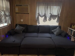 A sectional sofa with a remote control on it, a window with sheer curtains, and a wooden wall.