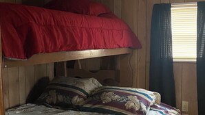 Ein Etagenbett mit rotem oberen Bett und einem unteren Bett mit Blumenmuster. Über dem Bett befindet sich ein Deckenventilator und zur Rechten ein Fenster mit Vorhängen.