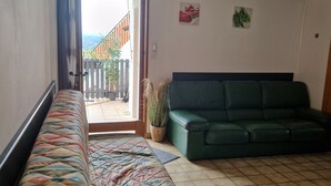 A room with a wooden bed, a green sofa, a tiled floor, and a view of a balcony with plants and a building. - Gîte de Pigneux (Saint-Genix-les-Villages)