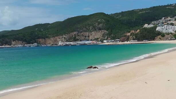 Una playa con aguas turquesas cristalinas, una orilla de arena y colinas verdes al fondo con edificios.