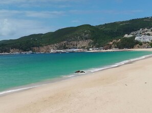 Una playa con aguas turquesas cristalinas, una orilla de arena y colinas verdes al fondo con edificios.