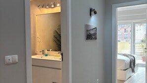 Un baño moderno con un lavamanos blanco, un espejo y una ducha. Hay una planta en maceta y un cuadro en la pared. El cuarto tiene paredes gris claro y piso de madera.