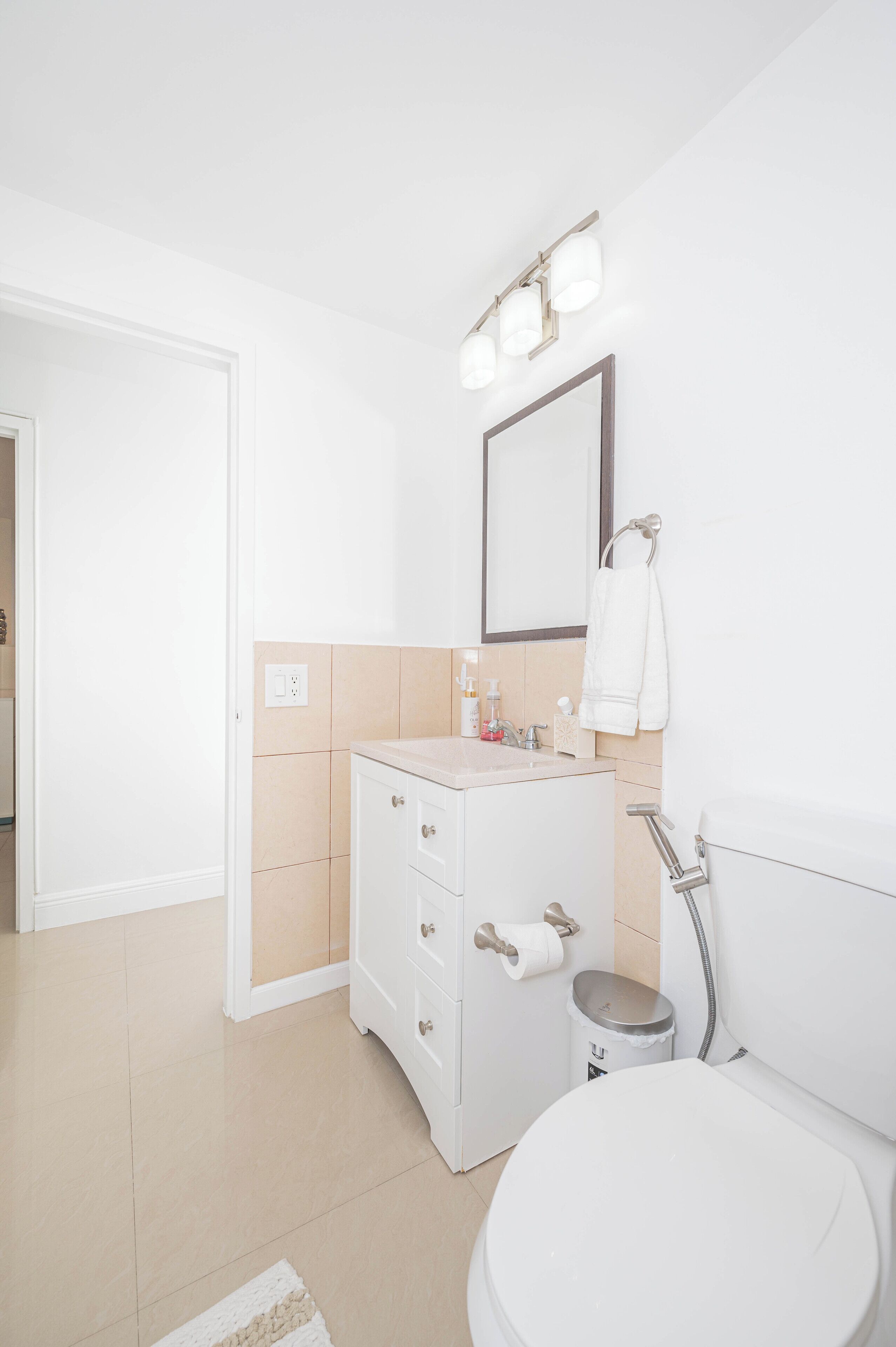 A modern bathroom with a white sink, mirror, and toilet. There is a towel rack above the sink and a trash can beside the toilet.