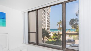 Une chambre d’hôtel avec une grande fenêtre donnant sur une plage, des palmiers et un immeuble imposant.