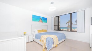 Una camera da letto con una grande finestra che si affaccia sulla spiaggia, un letto con copriletto a righe blu e bianche e un comodino bianco con un vaso di fiori.