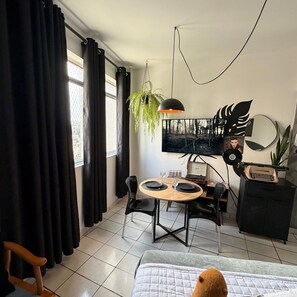 Ett modernt rum med en säng, ett matbord med stolar, ett litet skrivbord med en skrivmaskin, en hängande växt och en stor dekorativ spegel.