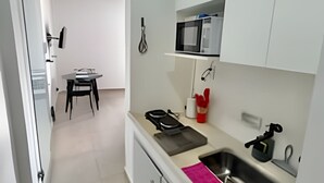A narrow kitchen with white cabinets, a microwave, a sink, and a dining area visible in the background. - KitiHaus Campinas (Campinas)