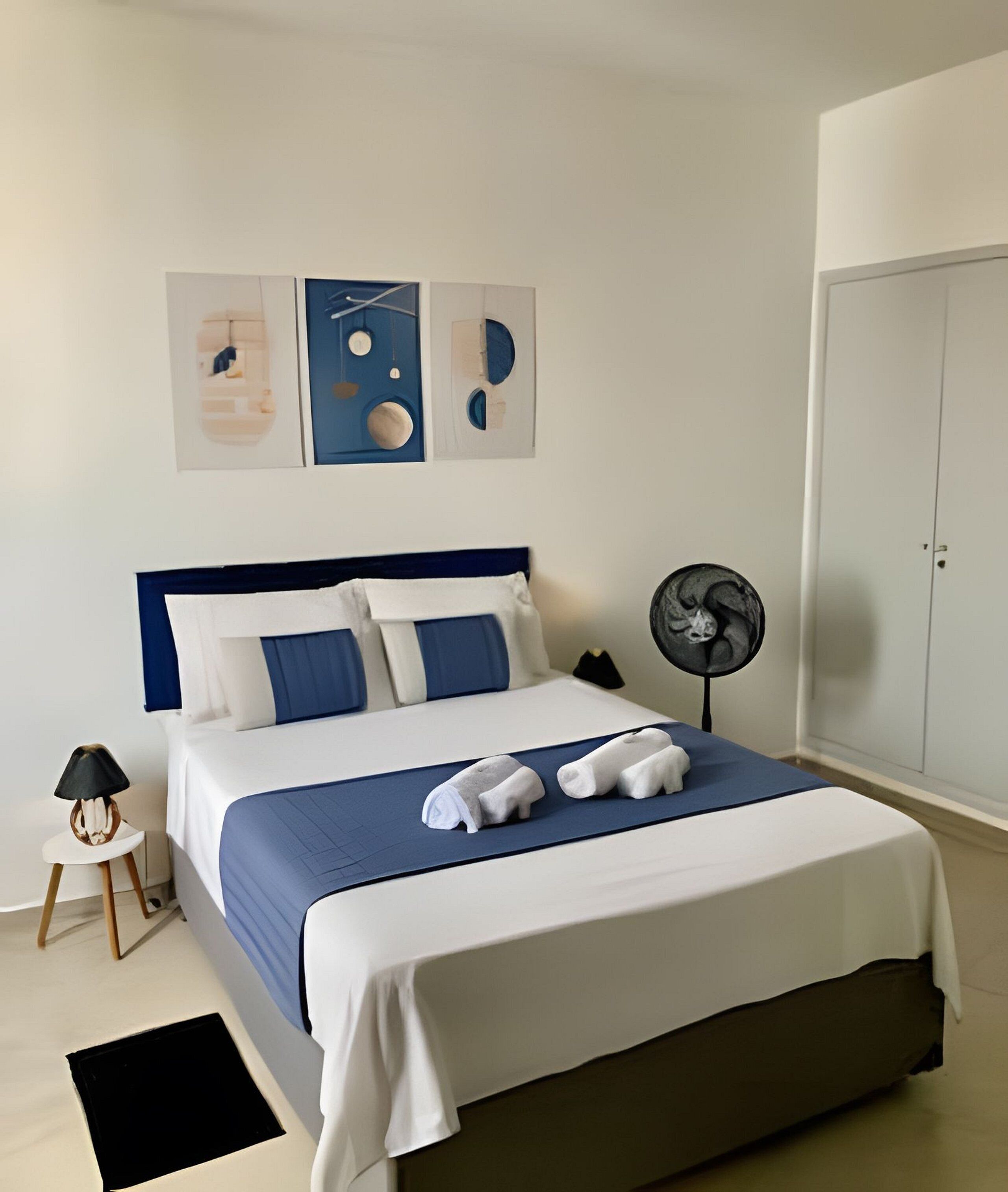 A neatly made bed with a blue and white color scheme, a bedside table with a lamp, and a wall-mounted fan.