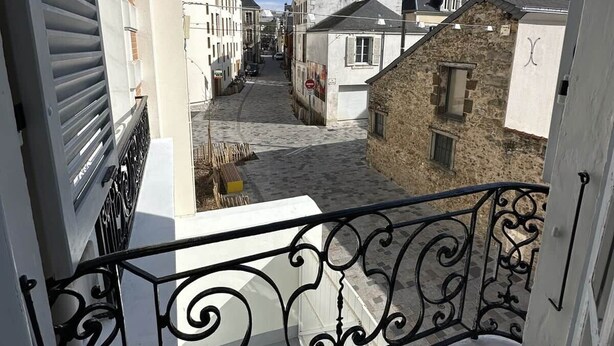 Una calle estrecha flanqueada por edificios, un cielo azul despejado y un balcón con barandillas de metal negras ornamentadas.