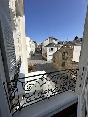 Une rue étroite bordée de bâtiments des deux côtés, un ciel bleu dégagé et un balcon orné de balustrades en métal noir ouvragé.