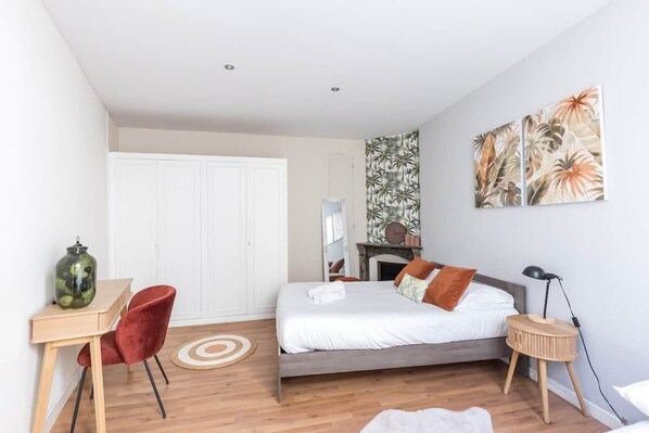 Une chambre avec un lit, un bureau, une chaise, un tapis rond, une table d’appoint, une lampe et un mur orné d’un motif représentant des plantes tropicales.