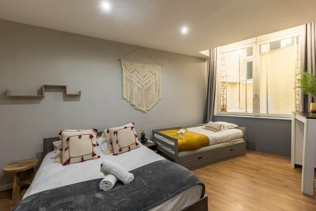 Une chambre moderne avec un lit, des tables de chevet, une décoration murale en macramé suspendue et une fenêtre avec des rideaux.