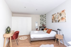Une chambre avec un lit, un bureau, une chaise, un tapis rond, une table d’appoint, une lampe et un mur orné d’un motif représentant des plantes tropicales.