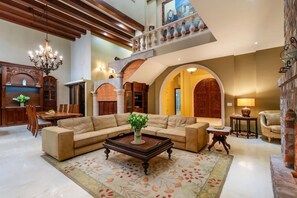 A spacious living area with a large sofa, a coffee table, and a chandelier. There is a balcony with a railing and a view of the outside. - Château Cerritos | Luxe Oceanfront Home (La Paz)