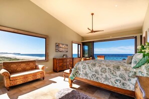 A bedroom with a large bed, a chair, a dresser, and a ceiling fan. It has large windows with a view of the beach.