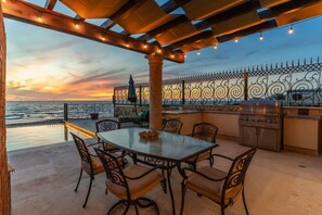 An outdoor patio with a table and chairs, a grill, and a view of the sea at sunset. - Château Cerritos | Luxe Oceanfront Estate (La Paz)
