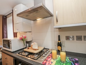 Une cuisine équipée d’un four à micro-ondes, d’un grille-pain, d’une planche à découper avec du pain, d’une cuisinière à gaz, d’une bouteille de Prosecco et de verres de jus d’orange.