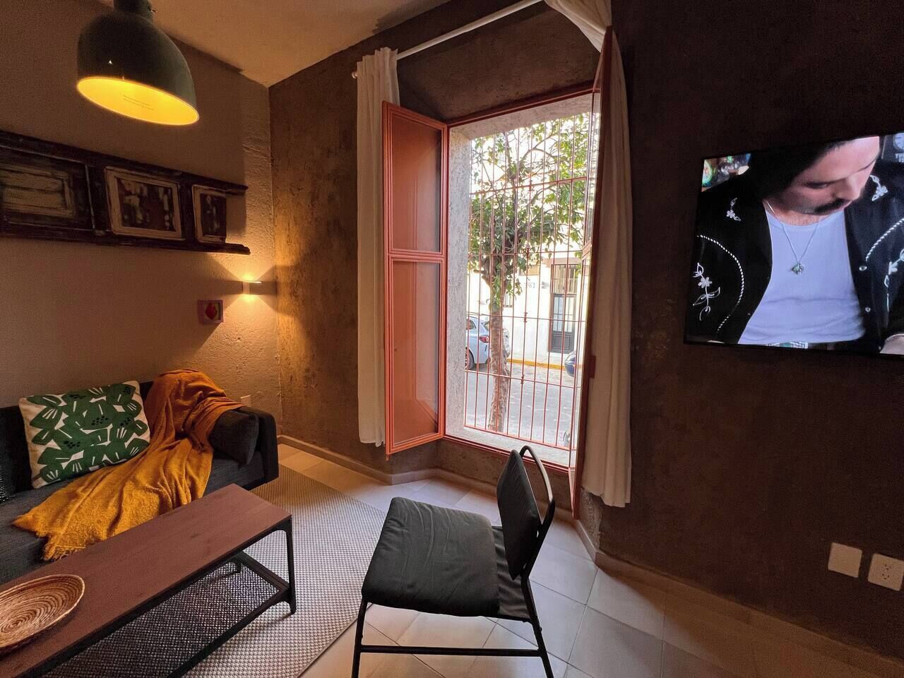 A living room with a sofa, a coffee table, a TV mounted on the wall, and a window with a view of the street.