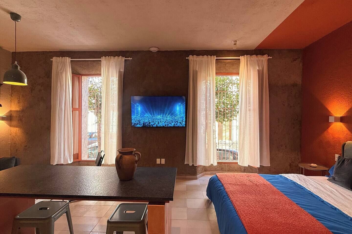 Una habitación con cama, televisor en la pared, mesa de comedor con sillas y un florero sobre la mesa.