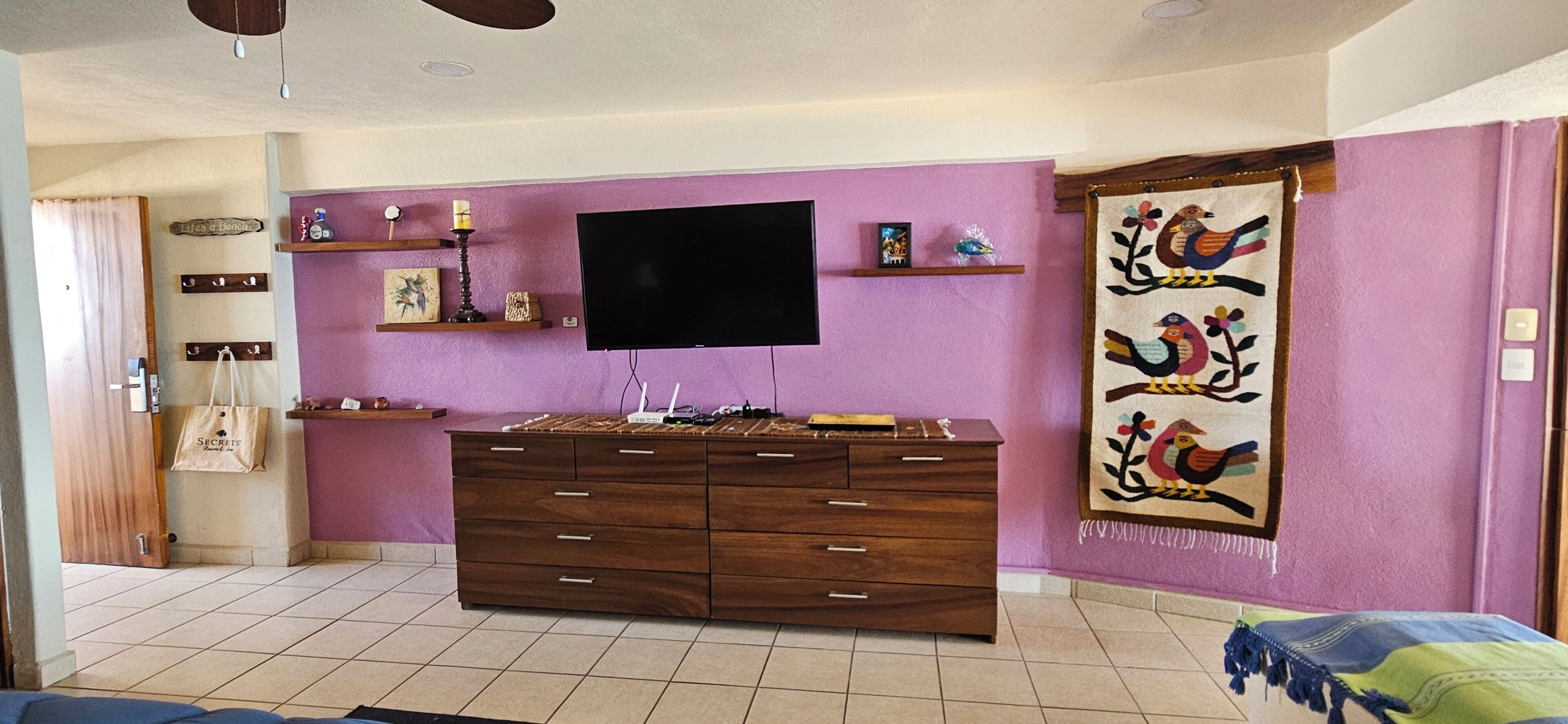 Ein Zimmer mit einem großen Flachbildfernseher an einer violetten Wand, einer Holzkommode mit Schubladen, einem Deckenventilator und einem Wandteppich mit Vogelmotiv.