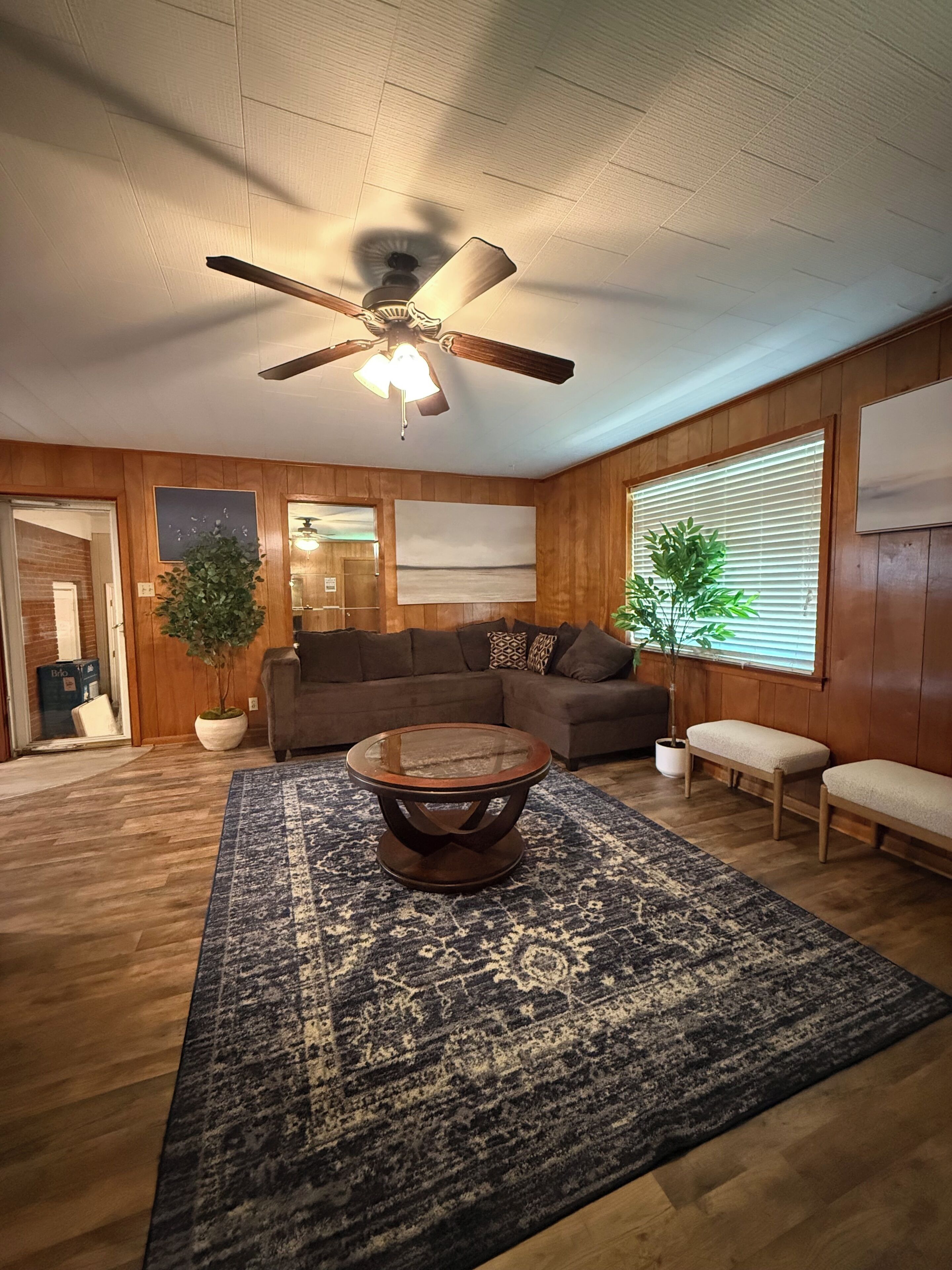A living room with a ceiling fan, a sectional sofa, a coffee table, and a large window with blinds.