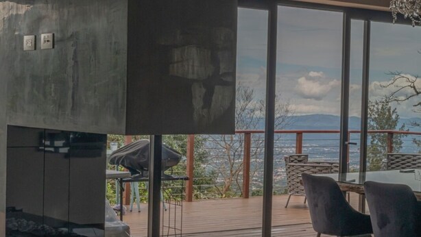 A modern interior with a stone fireplace, large windows offering a scenic view, and a chandelier. - Emerald Toucan Estate - 5 BR Mountain Retreat in Nature (San Bosco de Santa Barbara)