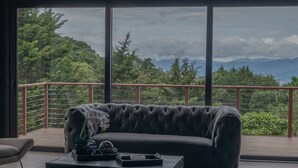 A modern living room with a dark leather sofa, a coffee table with decorative items, and a view of lush greenery and mountains through large windows. - Emerald Toucan Estate - 5 BR Mountain Retreat in Nature (San Bosco de Santa Barbara)