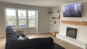 Ein Wohnzimmer mit einem Ecksofa, einem Kamin, einem Wand-Fernseher und einem großen Fenster.
